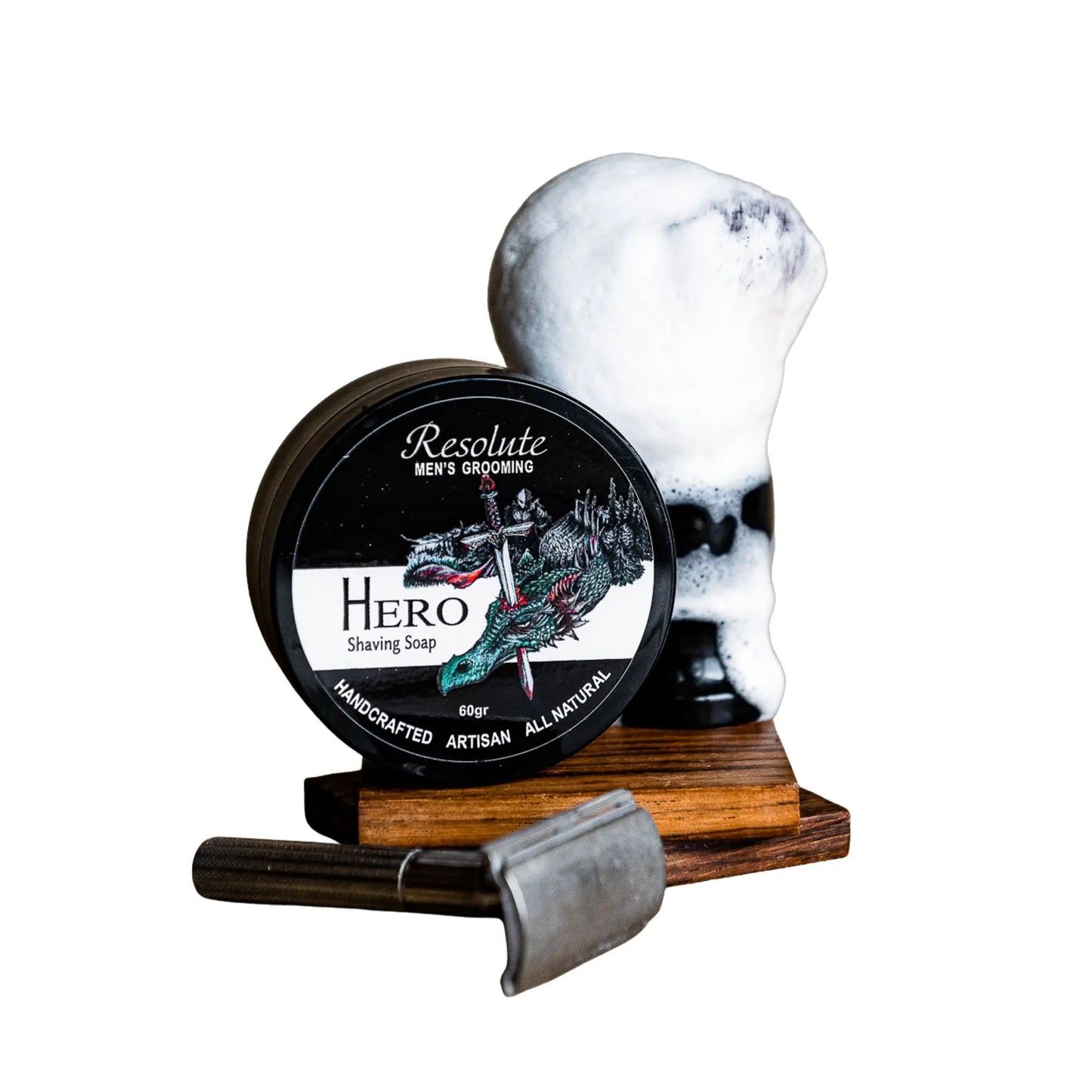 Resolute Hero Men's Grooming shaving soap in a round black tin with a dragon illustration, displayed next to a shaving foam head on a wooden stand with a metal razor in front, on a white background.
