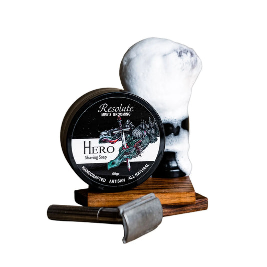 Resolute Hero Men's Grooming shaving soap in a round black tin with a dragon illustration, displayed next to a shaving foam head on a wooden stand with a metal razor in front, on a white background.