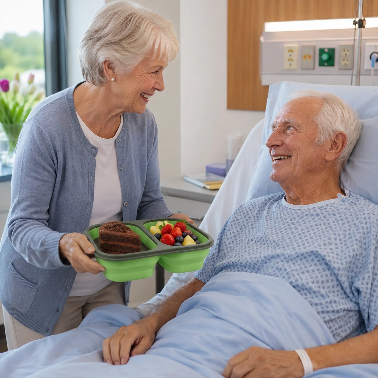 A smiling older woman in a hospital room holds a green two-compartment bento lunch tray with cake and fruit, while an older man lies in a hospital bed covered with a light blue blanket.