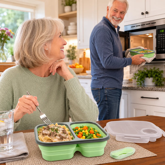 A woman sits at a kitchen table eating from a green compartment meal tray while a man in the background carries a container from a microwave. The meal tray contains pasta and mixed vegetables, with a clear lid and small accessory placed on the table.